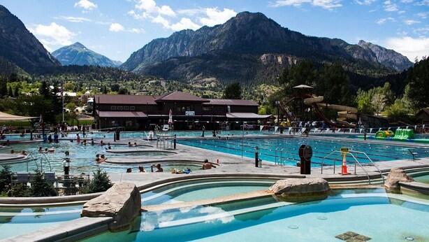 A pool area with multiple swimming pools, a building with a red roof, and mountains in the background. - Luxury Tiny Home/Base Camp to Ouray, Telluride/3 Queen beds/Washer & Dryer/Smart TV/Hot Tub Access/Patio w/Mountain Views/ Fire Pit/11 mi to Airport (Montrose)