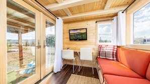 A cozy cabin interior with a wooden ceiling, a TV mounted on the wall, a sofa, and a dining area with two chairs. - Luxury Tiny Home/Base Camp to Ouray, Telluride/3 Queen beds/Washer & Dryer/Smart TV/Hot Tub Access/Patio w/Mountain Views/ Fire Pit/11 mi to Airport (Montrose)
