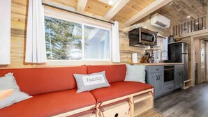 A cozy cabin interior with a red sofa, a kitchen area, and a window overlooking greenery. - Luxury Tiny Home/Base Camp to Ouray, Telluride/3 Queen beds/Washer & Dryer/Smart TV/Hot Tub Access/Patio w/Mountain Views/ Fire Pit/11 mi to Airport (Montrose)