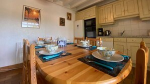 Zona pranzo con tavolo in legno apparecchiato per quattro persone, circondato da sedie. Si vede un angolo cottura con mobili chiari e un forno nero.