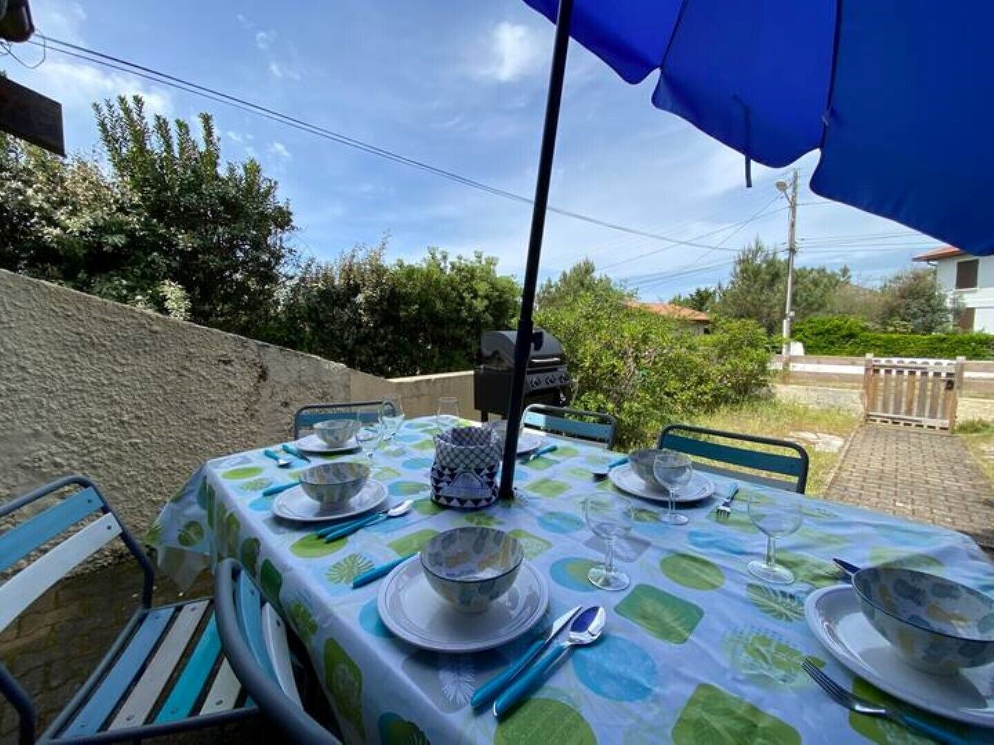En udendørs spiseplads med et bord dækket til fire personer, under en blå parasol, omgivet af grønt og en stenvæg.