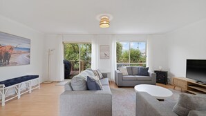 En moderne stue med en stor sofa, en mindre grå sofa, et sofabord og et fjernsyn. Der er et maleri af en tyr på væggen og et billede af en kænguru. Rummet har store vinduer med udsigt over grøn natur.