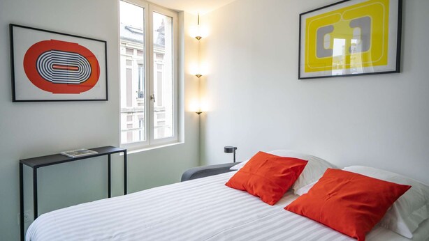 Een hotelkamer met een bed, twee kussens, een nachtkastje, een lamp en twee ingelijste kunstwerken aan de muur.