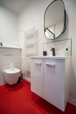 A modern bathroom with a white toilet, a white sink with a black faucet, a mirror, and a red patterned floor. - Alma (Centre-Ville - La Plage) (Le Havre)