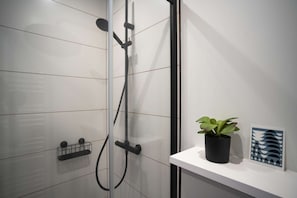 Un baño moderno con cabina de ducha de vidrio, un toallero negro, un pequeño estante con una planta en maceta y una obra de arte enmarcada.