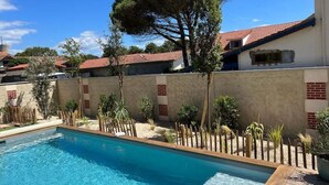 Una piscina con cubierta de madera, rodeada por un muro bajo con detalles en ladrillo rojo, y un árbol grande al fondo.