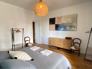 A bedroom with a bed, a dresser, a chair, a hanging light fixture, and a large abstract painting on the wall.