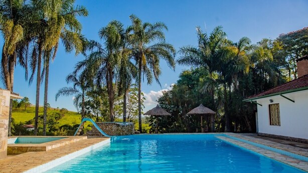 Una piscina con borde de piedra, rodeada de palmeras y un edificio blanco con techo de paja.