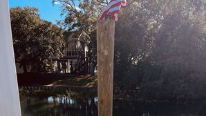 A wooden dock with a metal railing, a clear view of a calm body of water, and a building with a covered area in the background. - Love to fish! We have a dock  (Oak Island)