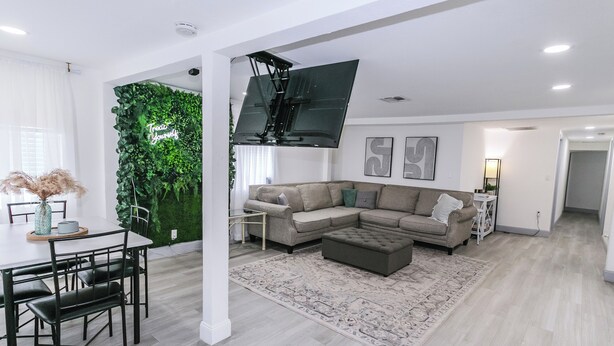 Una sala de estar moderna con un sofá modular, una mesa de comedor con sillas, un televisor de pantalla plana montado en una pared verde y una alfombra estampada.