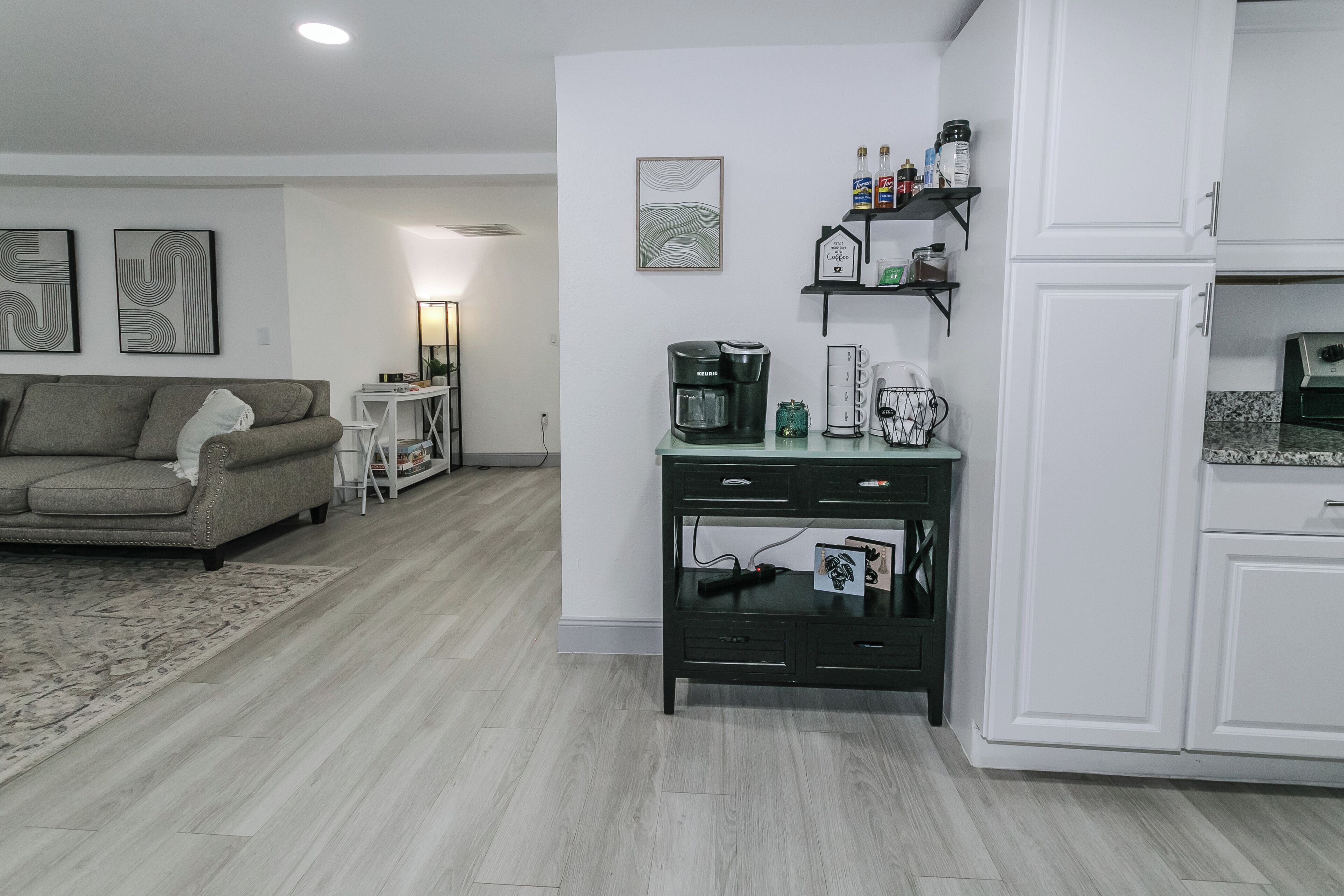A modern living room with a sofa, coffee table, and a small side table. There is a kitchen area with cabinets and a countertop.