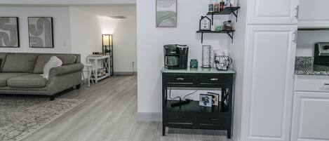 A modern living room with a sofa, coffee table, and a small side table. There is a kitchen area with cabinets and a countertop.