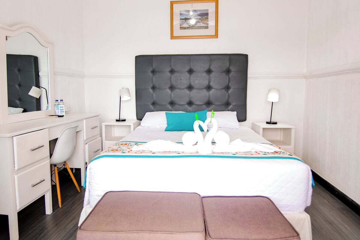 A hotel room with a bed, a desk, a chair, a ceiling fan, a bedside table, a lamp, a framed picture on the wall, and a window.