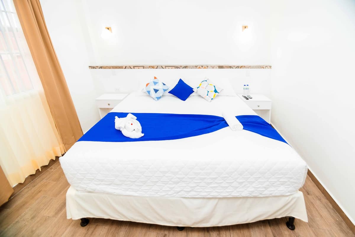 A neatly made bed with a blue bedspread and white bedding, two bedside tables with lamps, and a window with curtains.