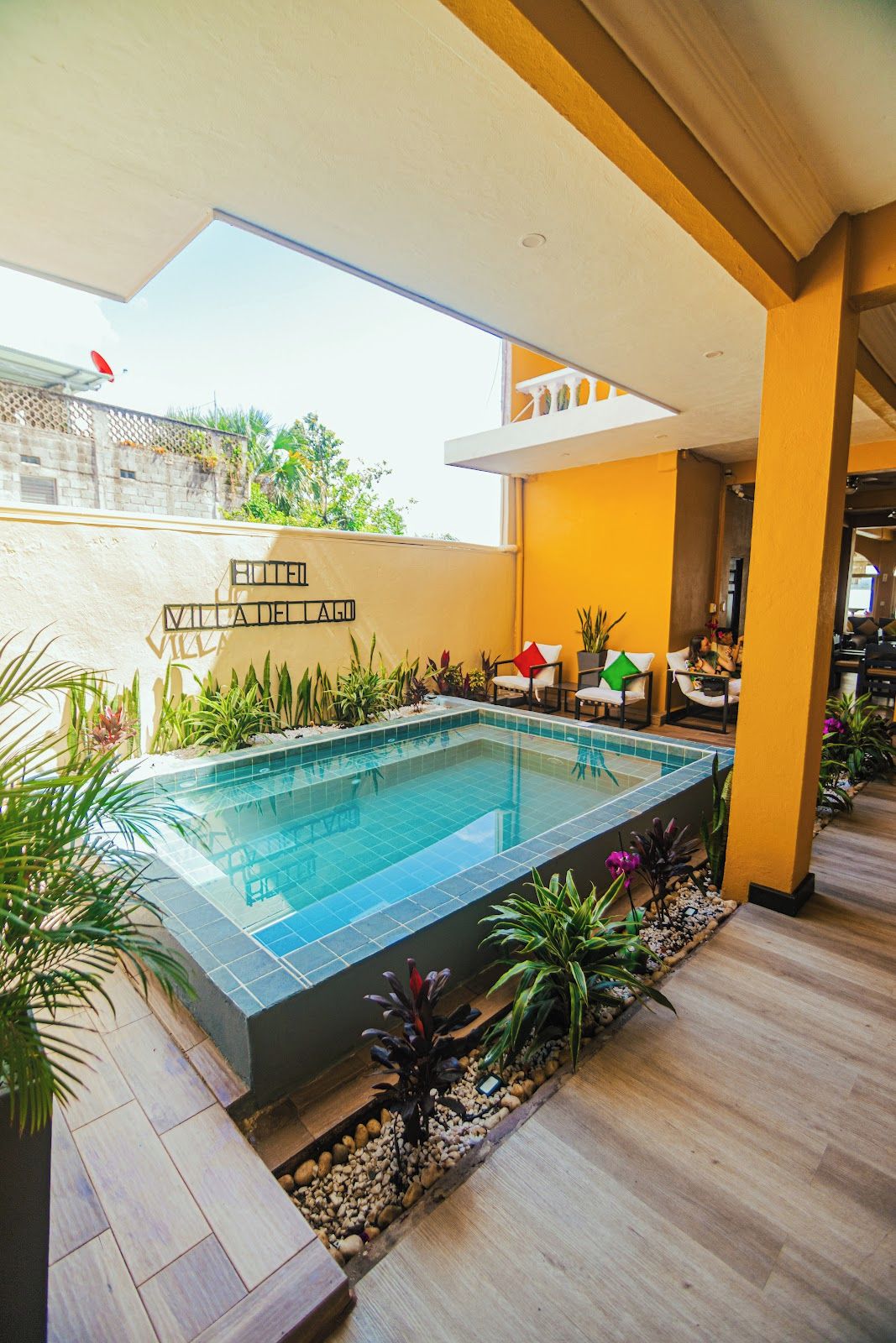 A swimming pool with a tiled deck, surrounded by plants and a small seating area with chairs.