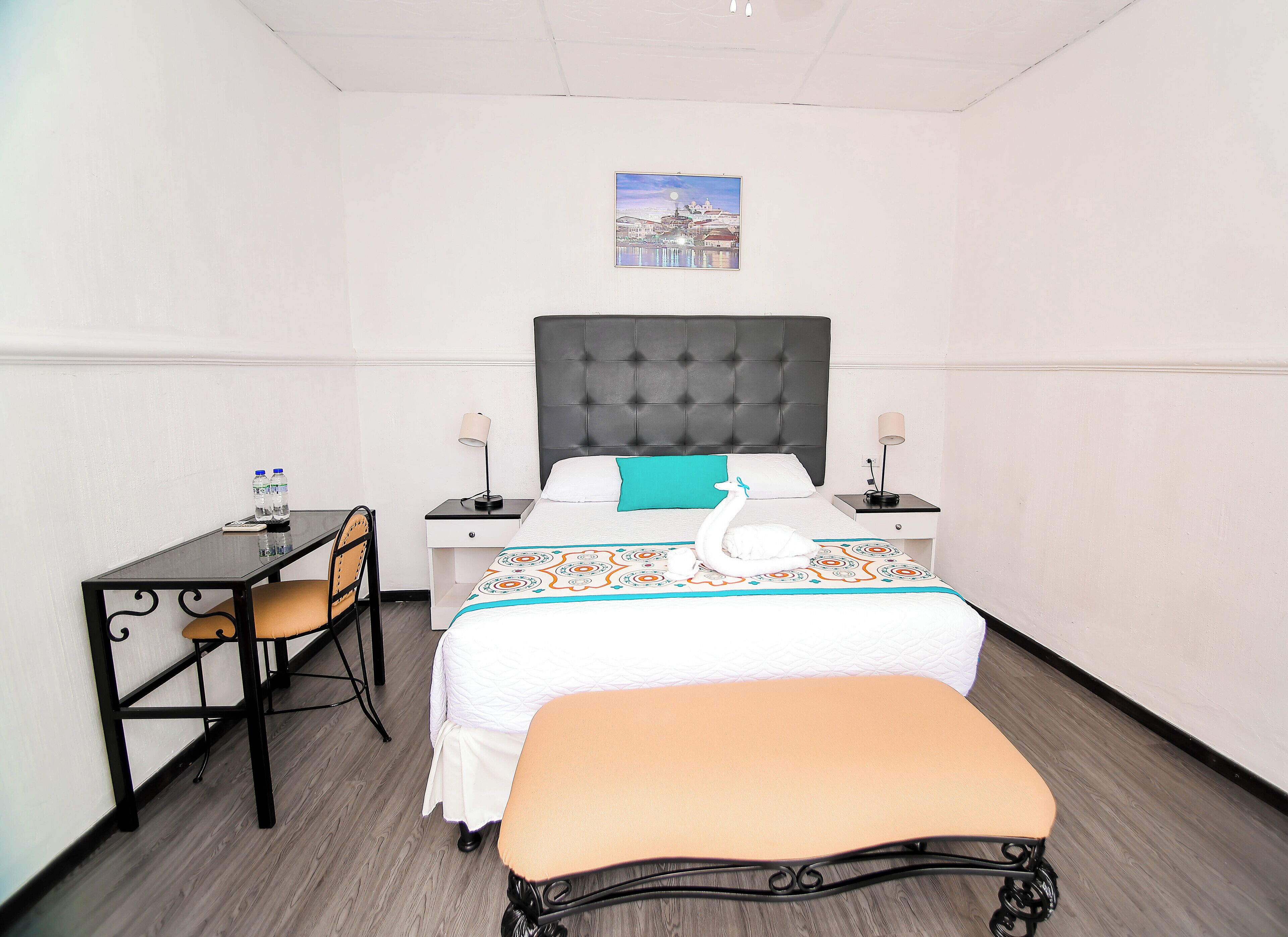 A hotel room with a bed, a desk, a chair, a bench, two bedside tables with lamps, and a wall-mounted picture.