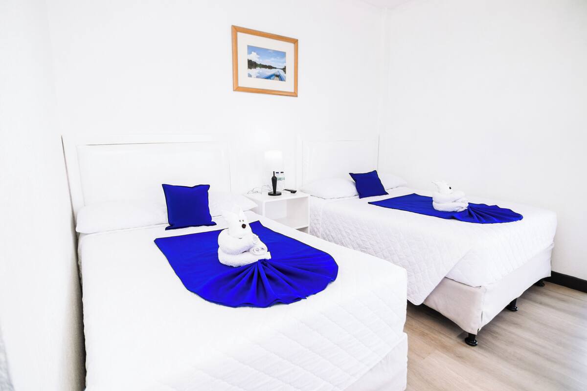 Two single beds with white bedding and blue accents, a small bedside table with a lamp, and a framed picture on the wall.