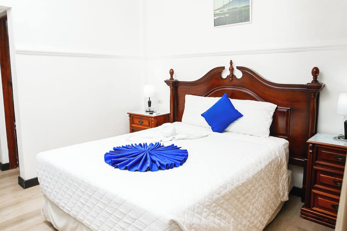 A neatly made bed with a white quilt and a blue decorative item, a wooden nightstand with a lamp, and a framed picture on the wall.