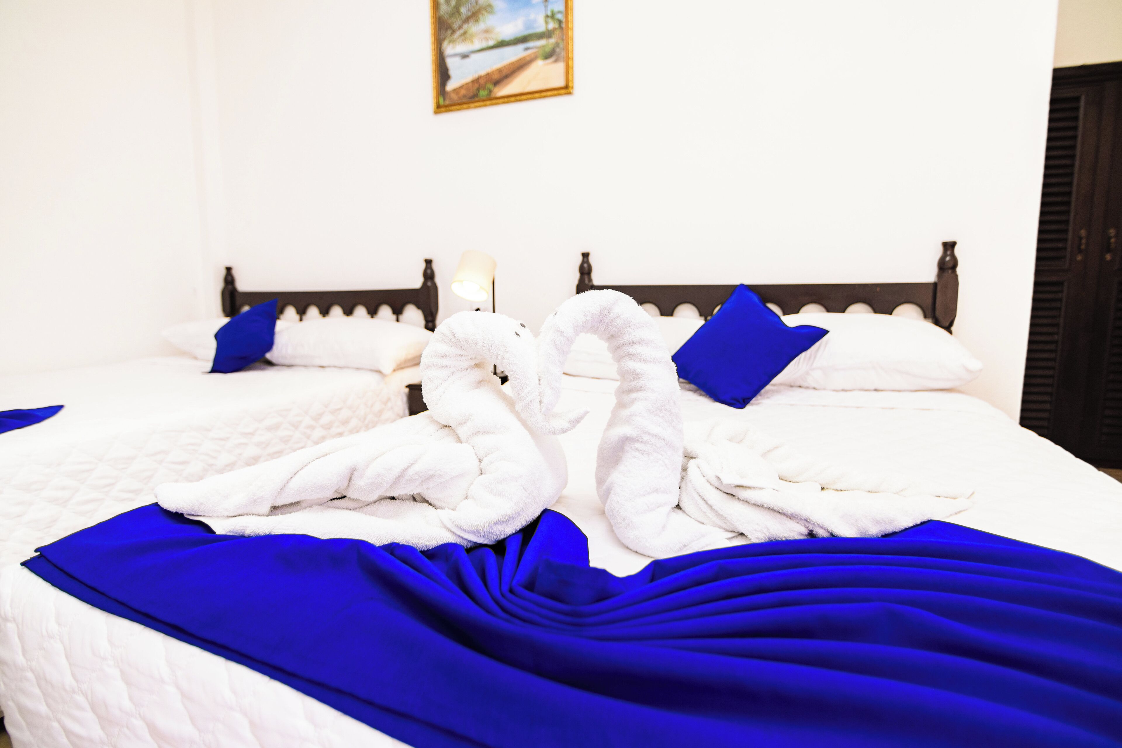 Een net opgemaakt bed met witte lakens, blauwe kussens en een blauwe deken, en een schilderij aan de muur.