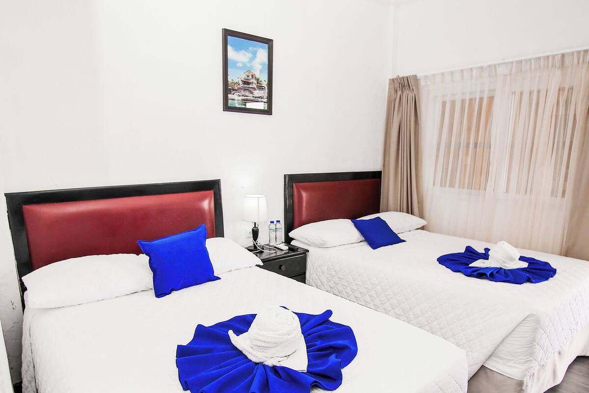A hotel room with two beds, each with a red headboard, white bedding, and blue decorative pillows. There is a nightstand with a lamp and a framed picture on the wall.