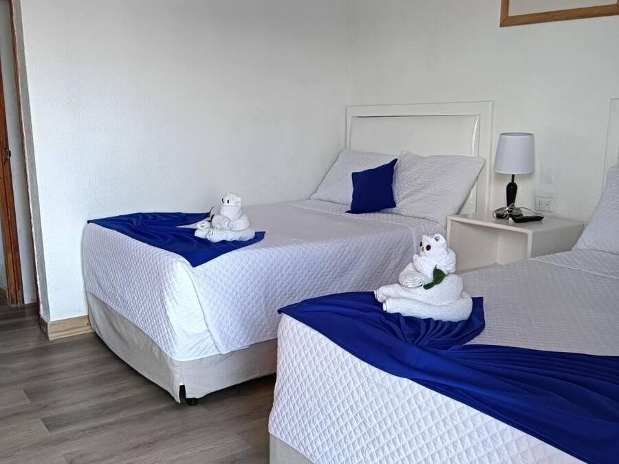 Dos camas individuales con ropa de cama blanca y detalles azules, una mesita de noche blanca con lámpara, y un cuadro en la pared.