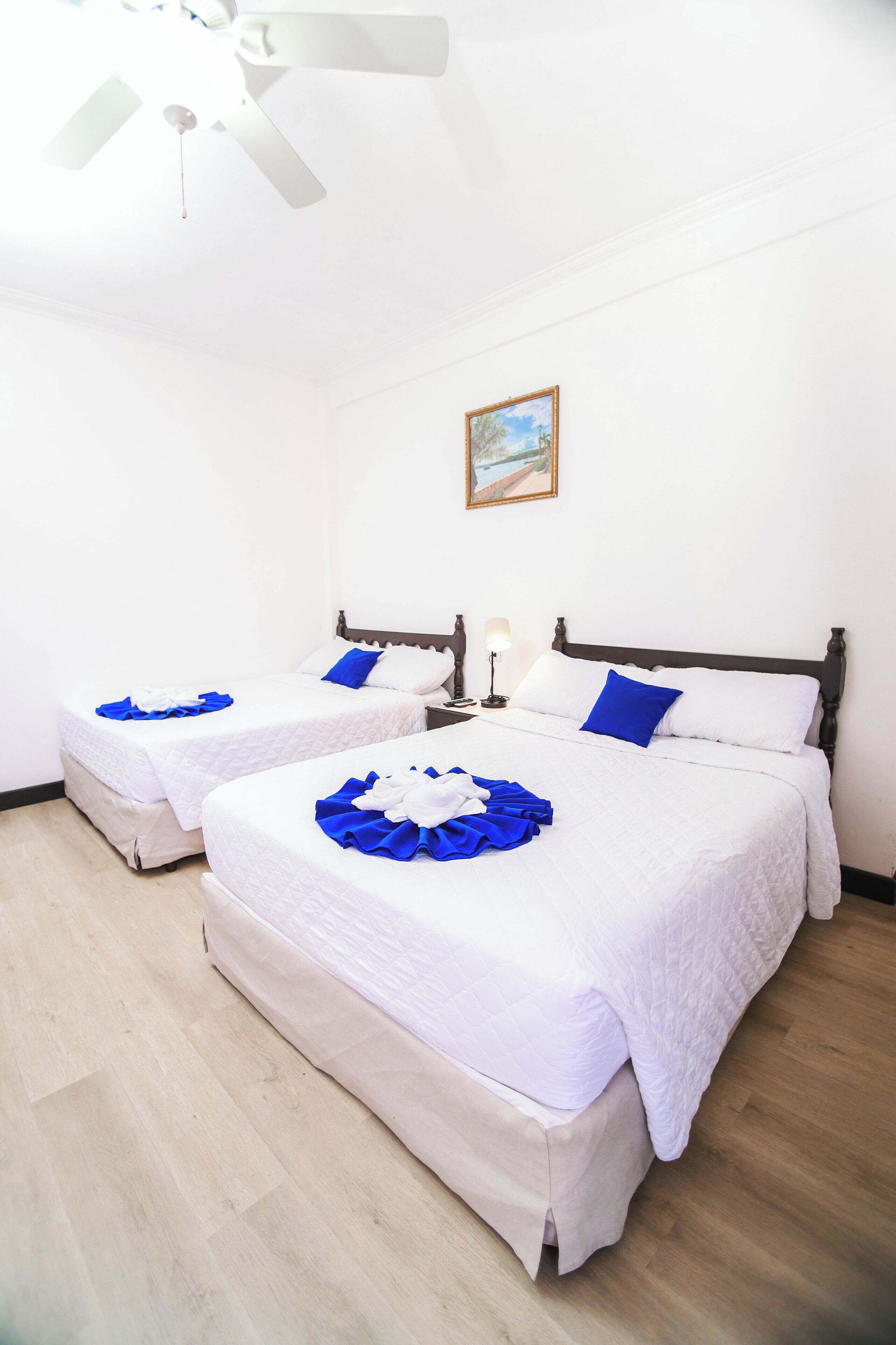 Habitación de hotel con dos camas, ropa de cama blanca, ventilador de techo y un cuadro en la pared.