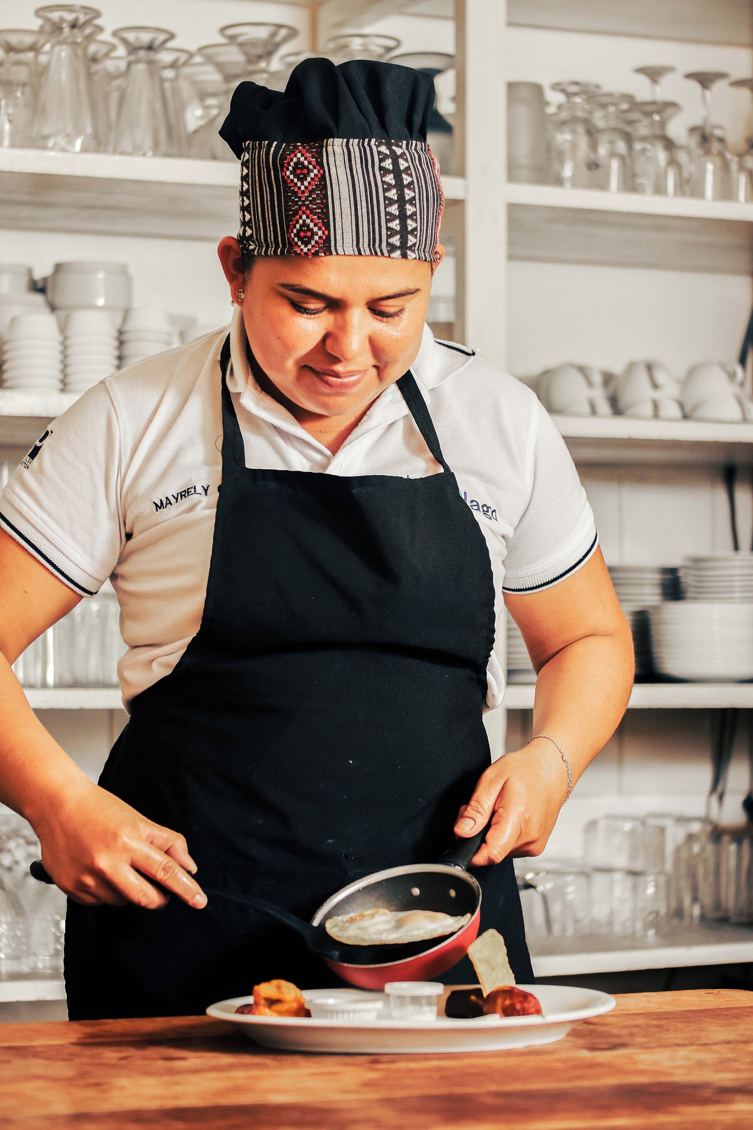 A person in a chef's uniform preparing food in a kitchen with shelves of glassware in the background. - Hotel Villa del Lago (Flores)