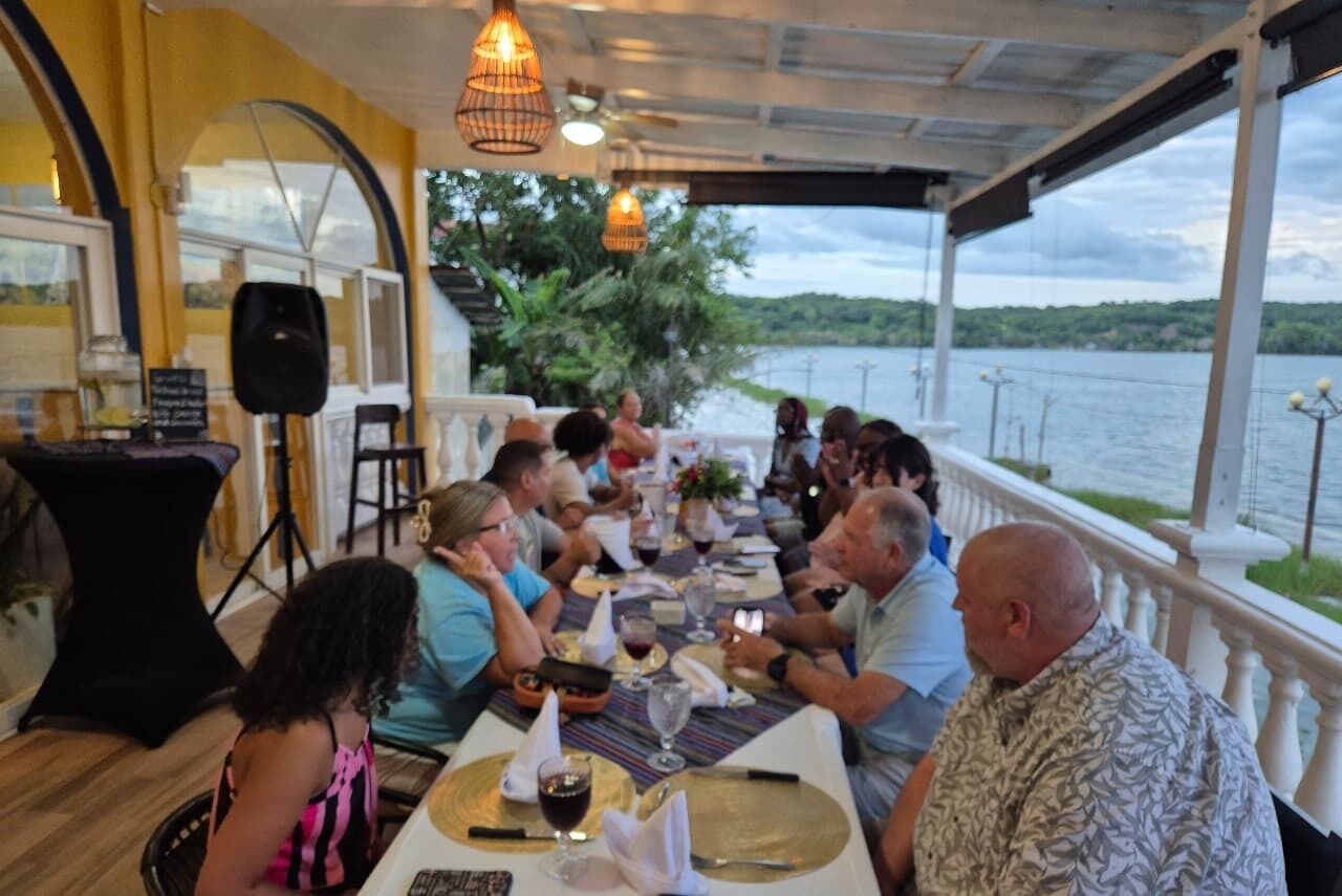 Un groupe de personnes est assis à une table en plein air, près d’un plan d’eau, et y prend son repas.