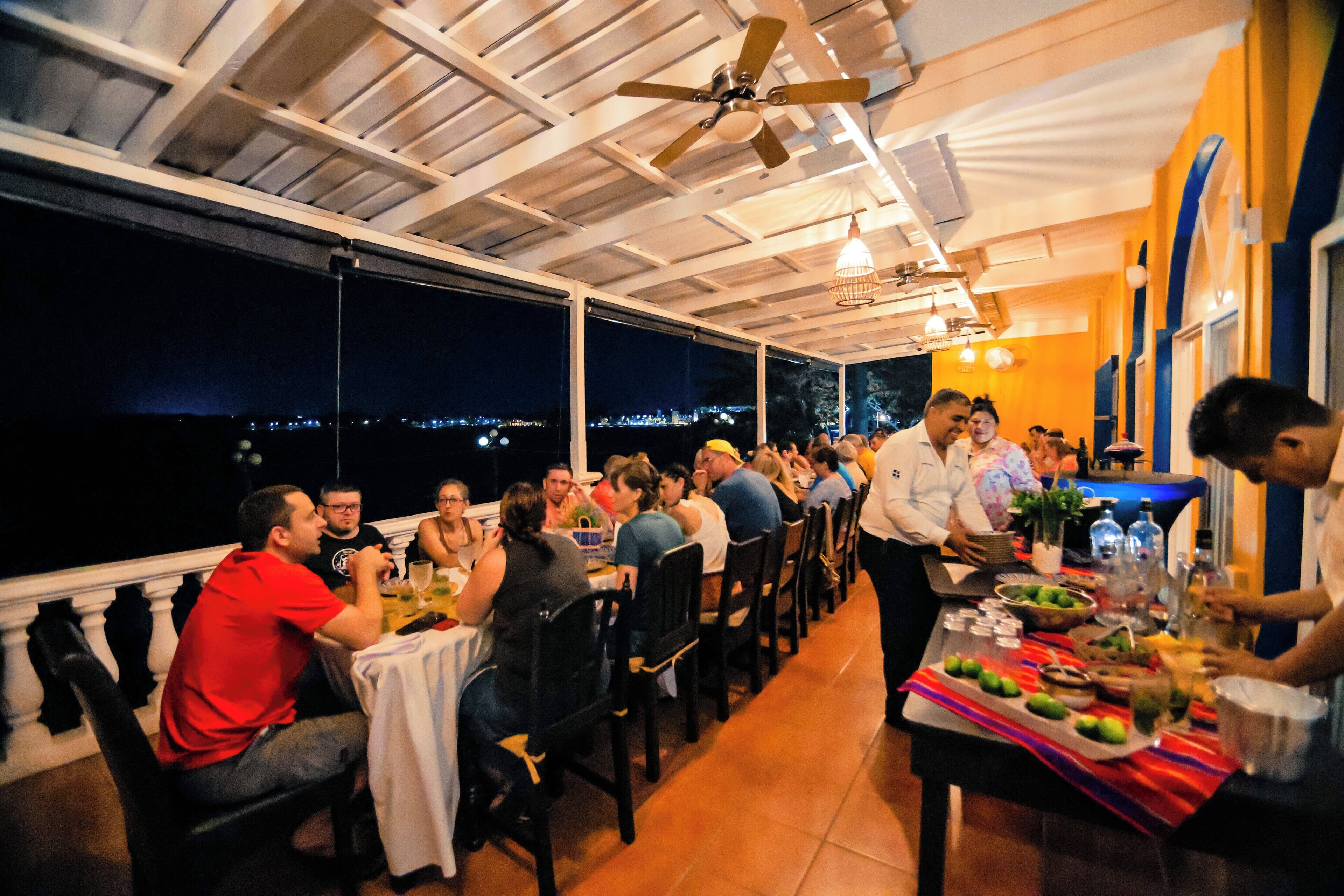 Un espace extérieur abrité, aménagé pour les repas, avec des tables et des chaises, un barman préparant des boissons et une vue sur la ville la nuit.
