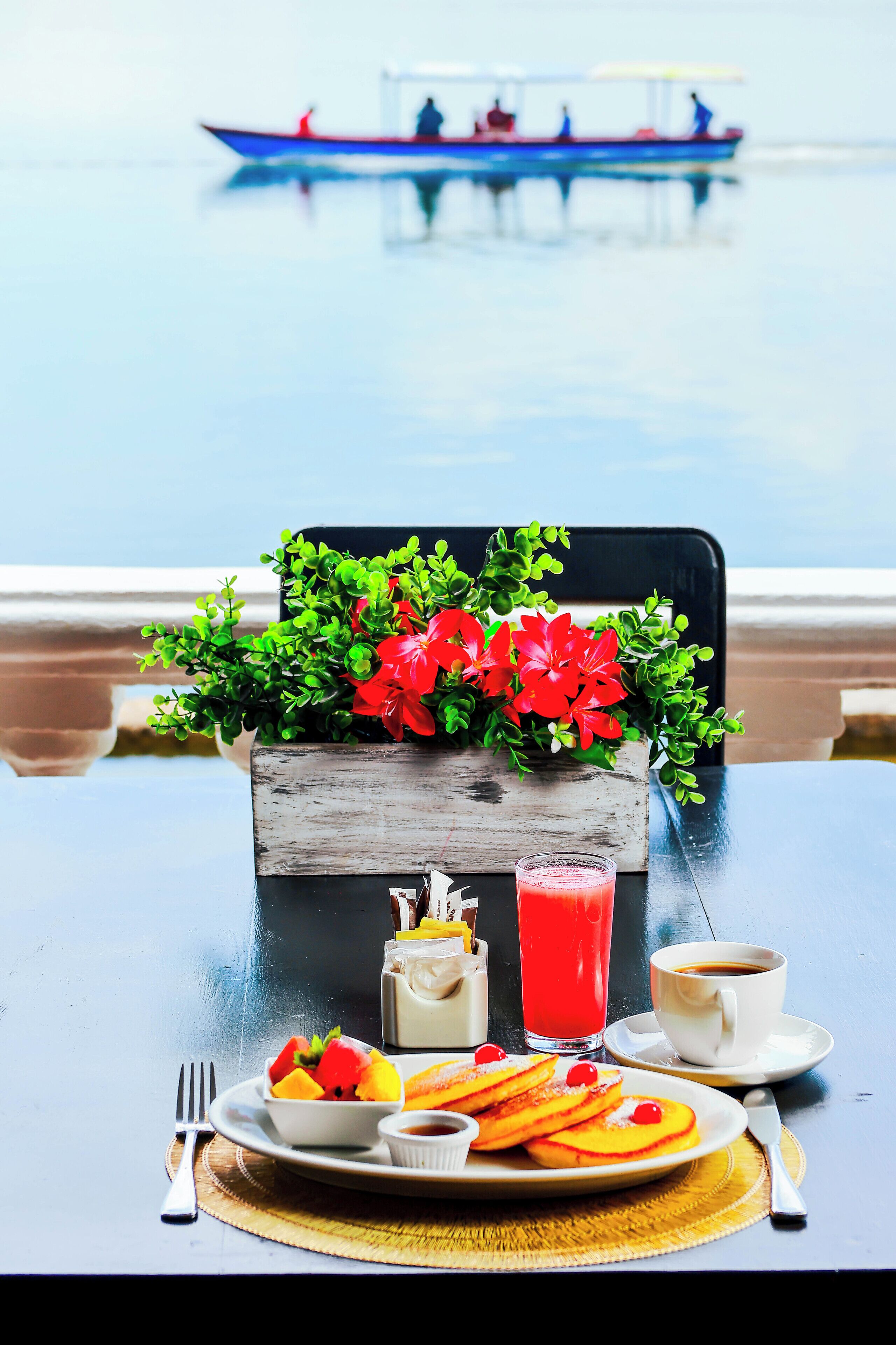 A table with breakfast items, a flower arrangement, and a view of a boat on water.