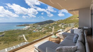 Een balkon met uitzicht op zee en heuvels, een bank en een tafel met een fles.