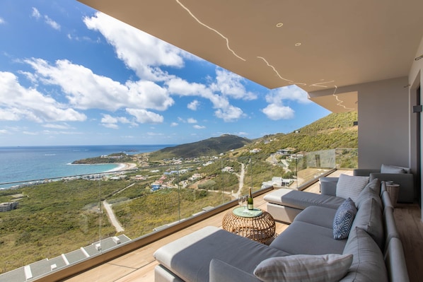A balcony with a view of the sea and hills, a sofa, and a table with a bottle.