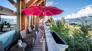 Una terraza de madera con muebles de mimbre y un paraguas rojo, con vistas a un paisaje montañoso.