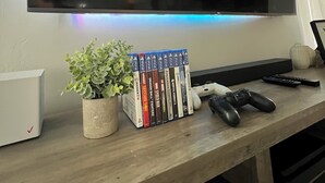 A flat-screen TV mounted on a wall, a gaming console, a plant, and a row of video game cases on a wooden desk. - Spacious 4-bedroom house in charming Carlsbad with WiFi and AC (Carlsbad)