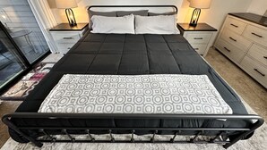 Una cama bien hecha, con estructura negra y colcha estampada, flanqueada por dos mesitas de noche con lámparas, un aparador y vistas a un balcón.