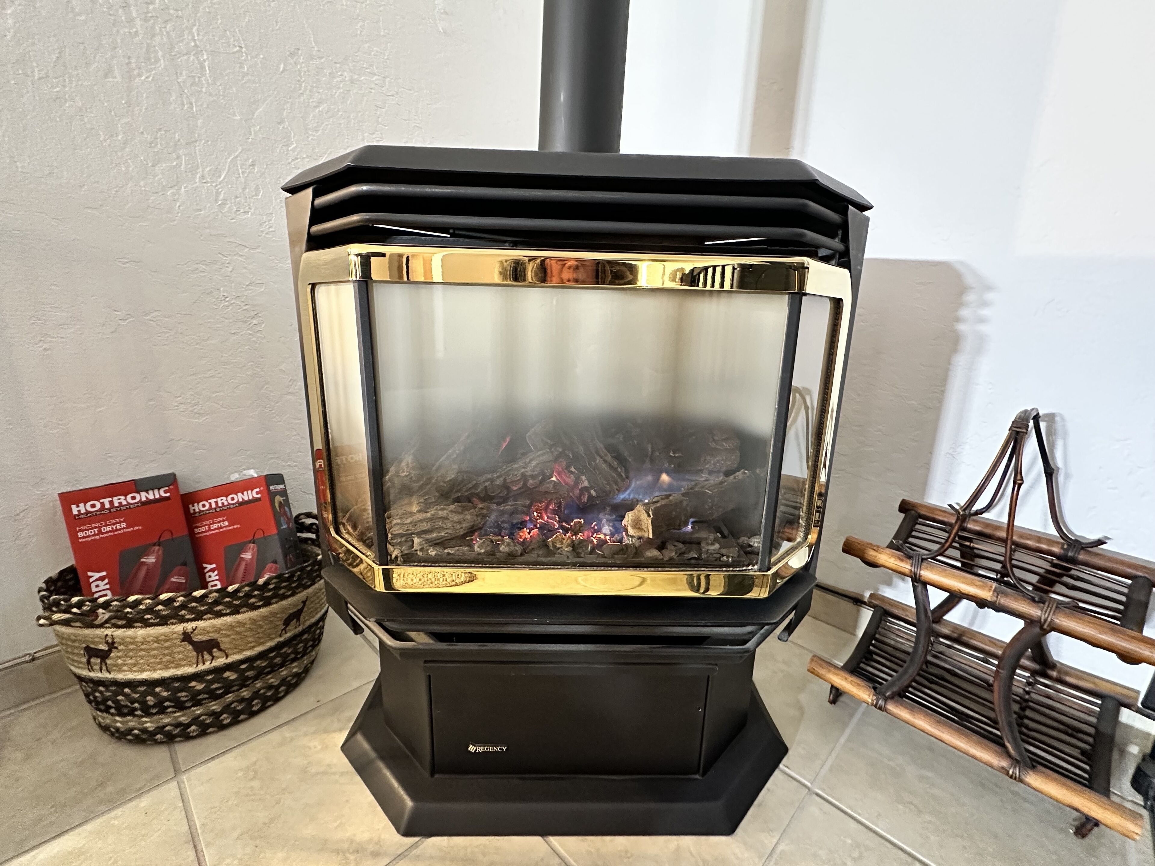 A wood-burning stove with a glass front, a black base, and a chimney. There are two boxes labeled "HOTTRONIC" and a woven basket with a wooden rack beside it.