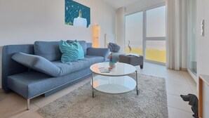 A modern living room with a blue sofa, a glass coffee table, and a large window offering a view of the outdoors. - Düne 1, Ferienwohnung 11 (Cuxhaven)