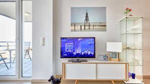 A modern living room with a flat-screen TV, a glass cabinet, and a painting of a lighthouse. - Düne 1, Ferienwohnung 11 (Cuxhaven)