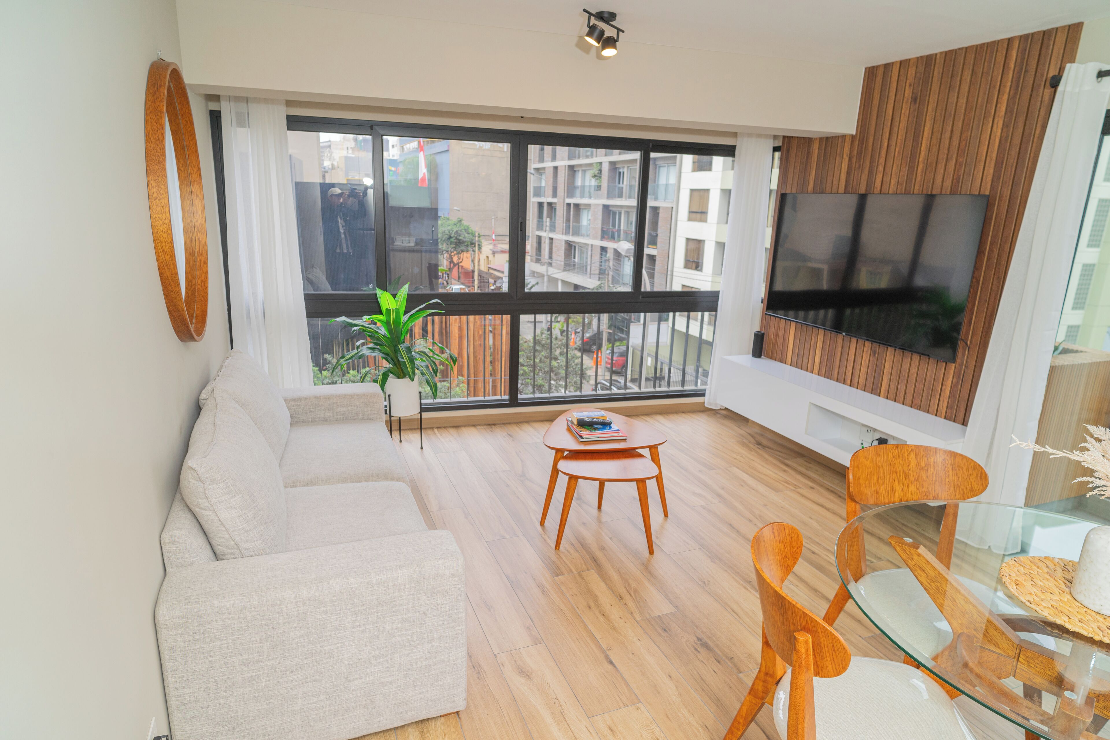 A modern living room with a sofa, a glass table, wooden chairs, a TV, and a large window with a view of buildings.