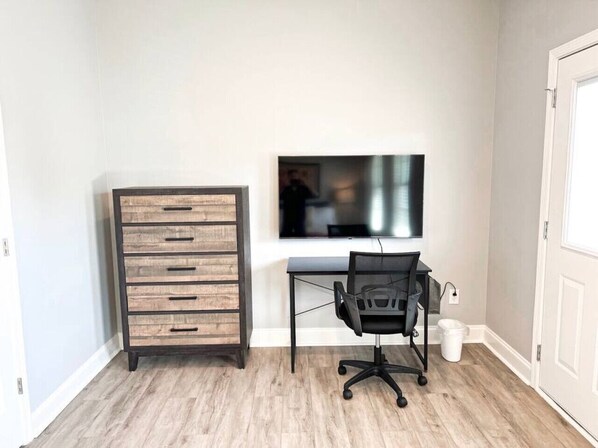 Ein Zimmer mit einer hölzernen Kommode, einem schwarzen Bürostuhl, einem Schreibtisch und einem an der Wand befestigten Fernseher.