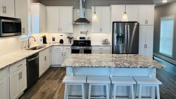 A modern kitchen with white cabinets, a granite island, stainless steel appliances, and pendant lighting.