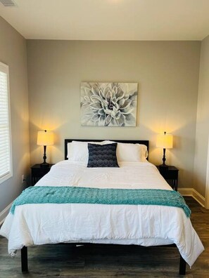 Un lit bien fait, recouvert d’un plaid turquoise, avec deux lampes de chevet et une œuvre d’art florale au-dessus du lit, dans une pièce dotée d’un plancher en bois et d’une fenêtre sur le côté gauche.