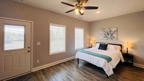 A bedroom with a bed, a ceiling fan, two windows with blinds, a door, and a wall-mounted artwork.