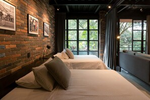 Een kamer met een bakstenen muur, twee bedden met kussens, een raam met gordijnen en ingelijste foto's aan de wand.