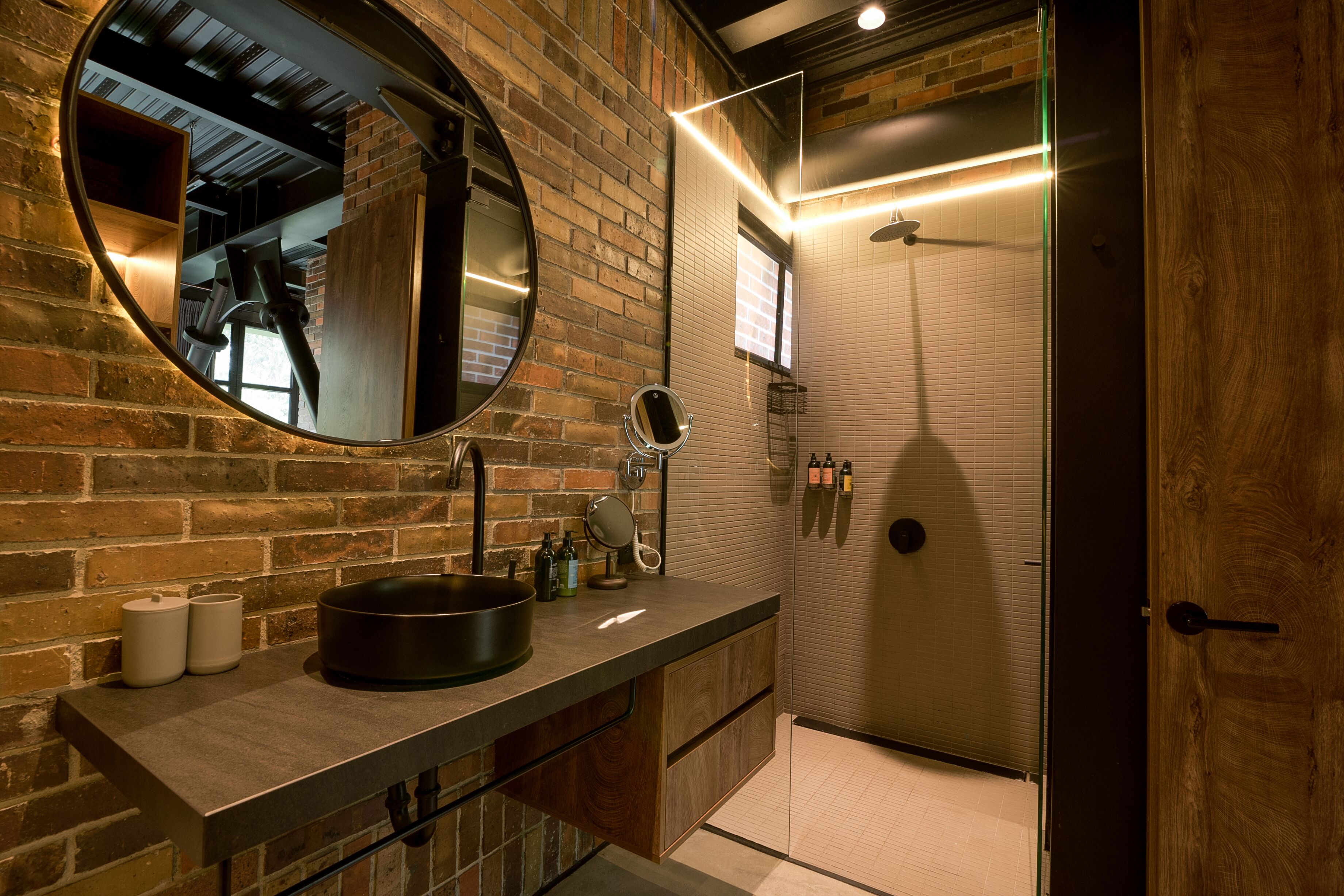 A modern bathroom with a brick wall, a round mirror, a sink with a black basin, and a shower area.