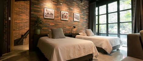 Habitación de hotel con dos camas, una pared de ladrillo y ventanas grandes.