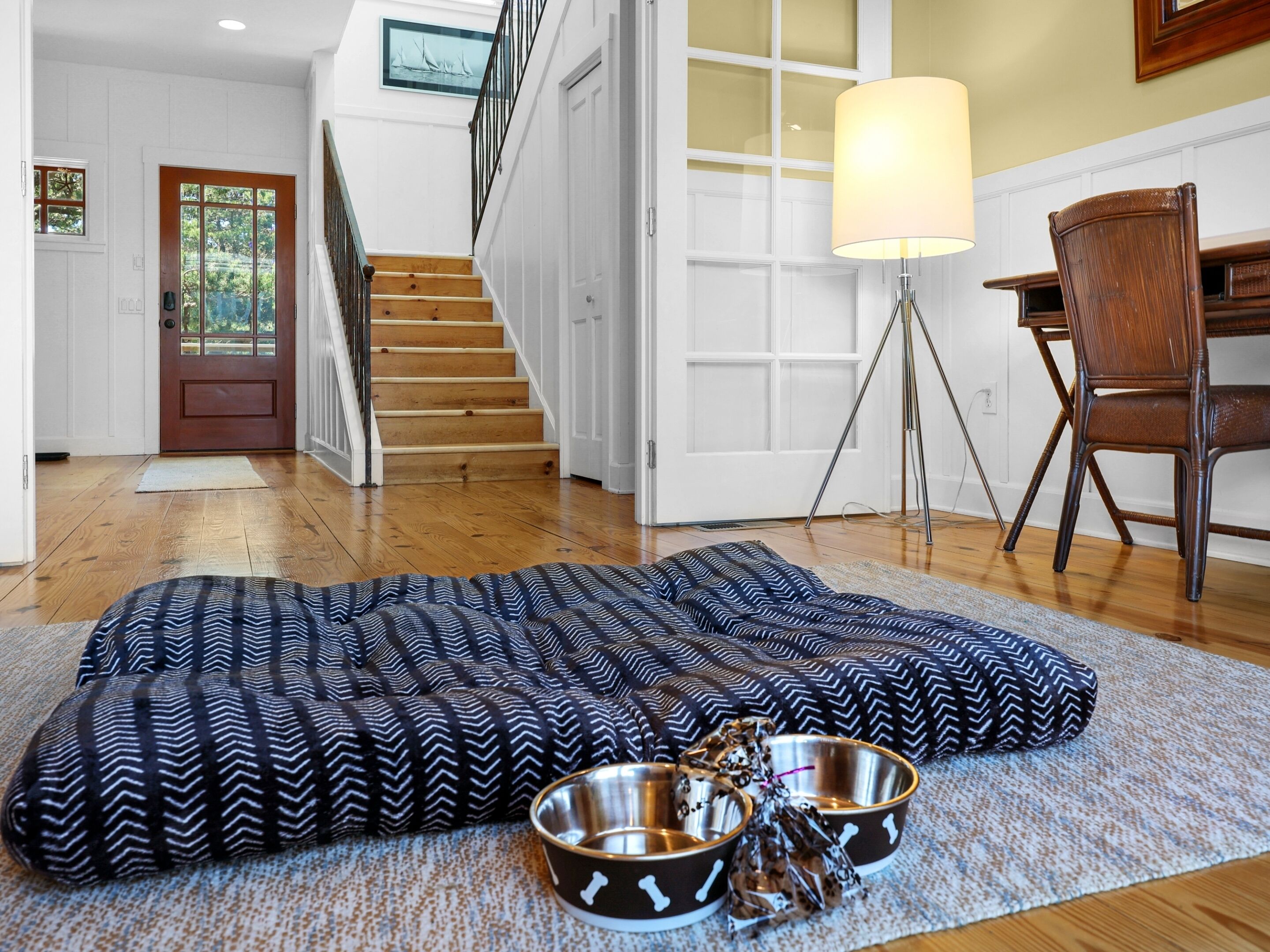 A room with a wooden floor, a staircase, a door with glass panels, a desk with a lamp, and two pet bowls on a patterned rug.