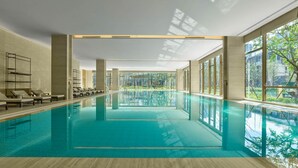 Une grande piscine intérieure aux parois en verre transparent, dotée de chaises longues et offrant une vue sur la verdure environnante.