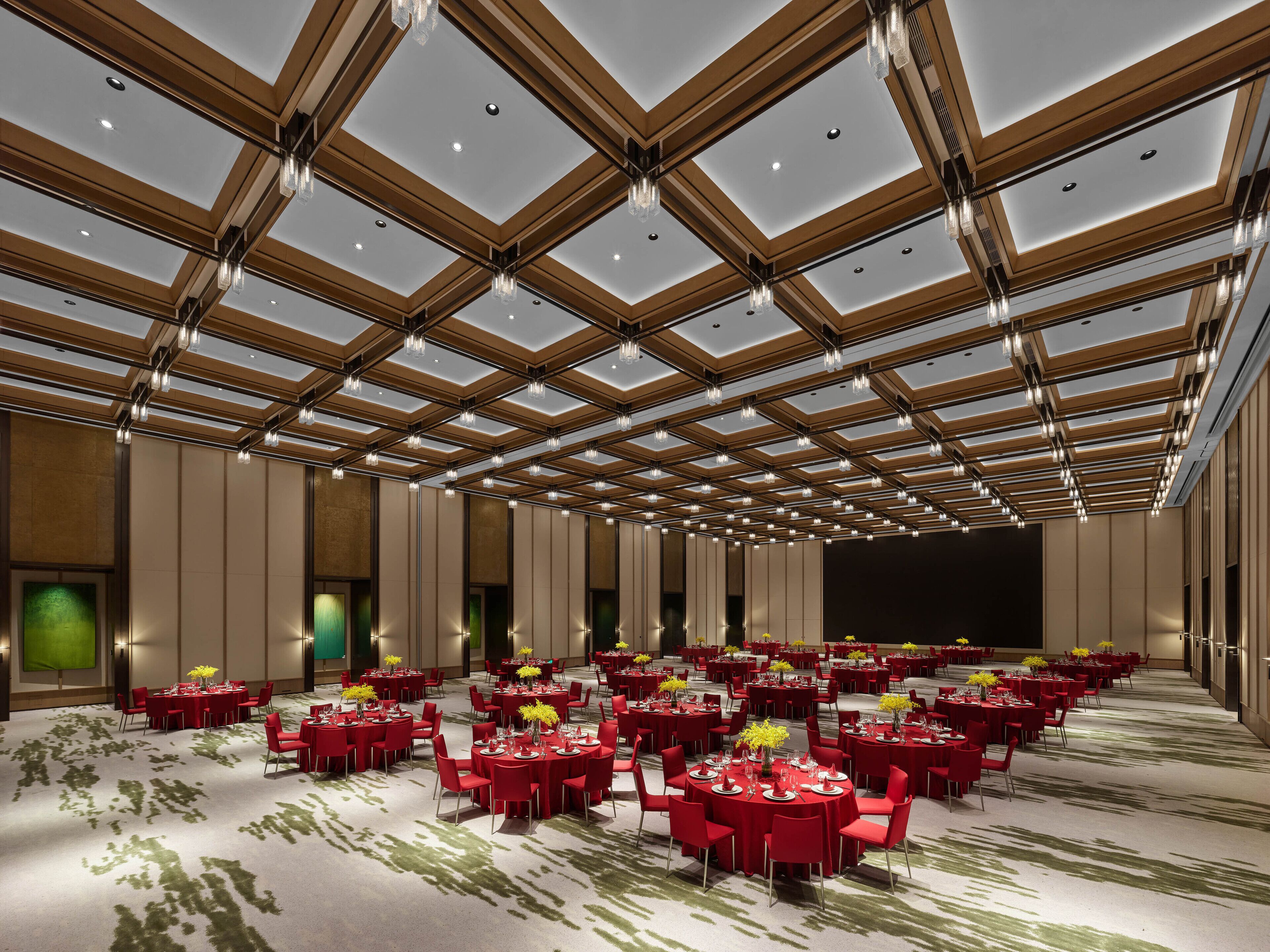 Une vaste salle de banquet dotée de nombreuses tables rondes, chacune recouverte d’une nappe rouge et agrémentée de chaises, et un plafond haut orné d’un motif géométrique en bois.