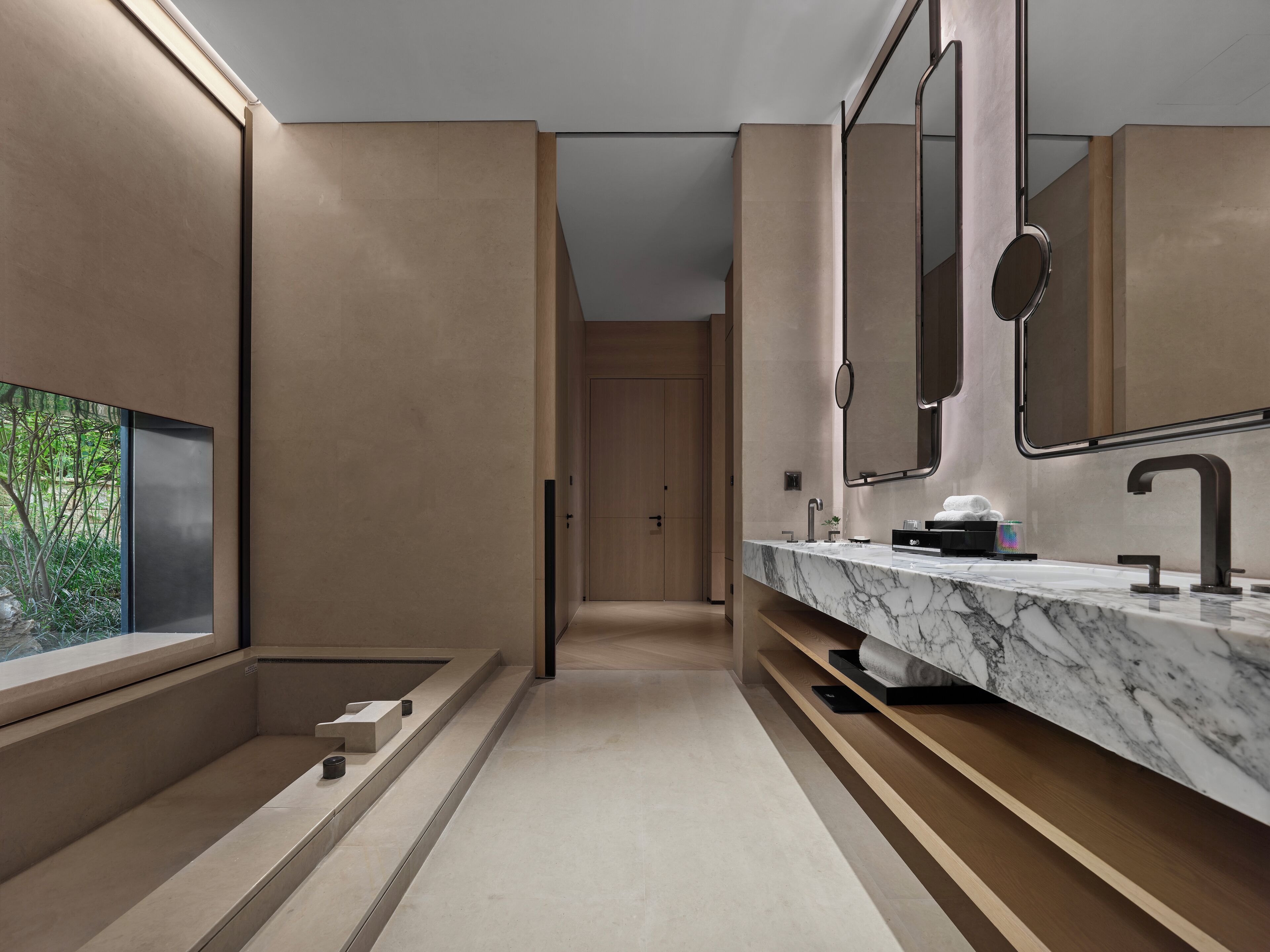 A modern bathroom with a marble sink, large mirrors, and a view of greenery through a window.